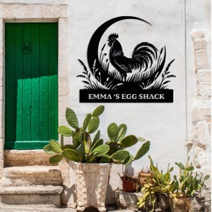 May include: Black metal rooster sign with the text "EMMA'S EGG SHACK" displayed on a white wall. The sign features a rooster silhouette within a crescent moon and grass design. A green door and potted plants are in the background.