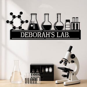 Könnte beinhalten: Schwarze Metall-Wandkunst mit Laborgeräten und einer Molekularstruktur. Das Schild lautet "DEBORAH'S LAB". Ein Mikroskop, Bechergläser und ein kleiner Schrank mit Fläschchen sind ebenfalls zu sehen.