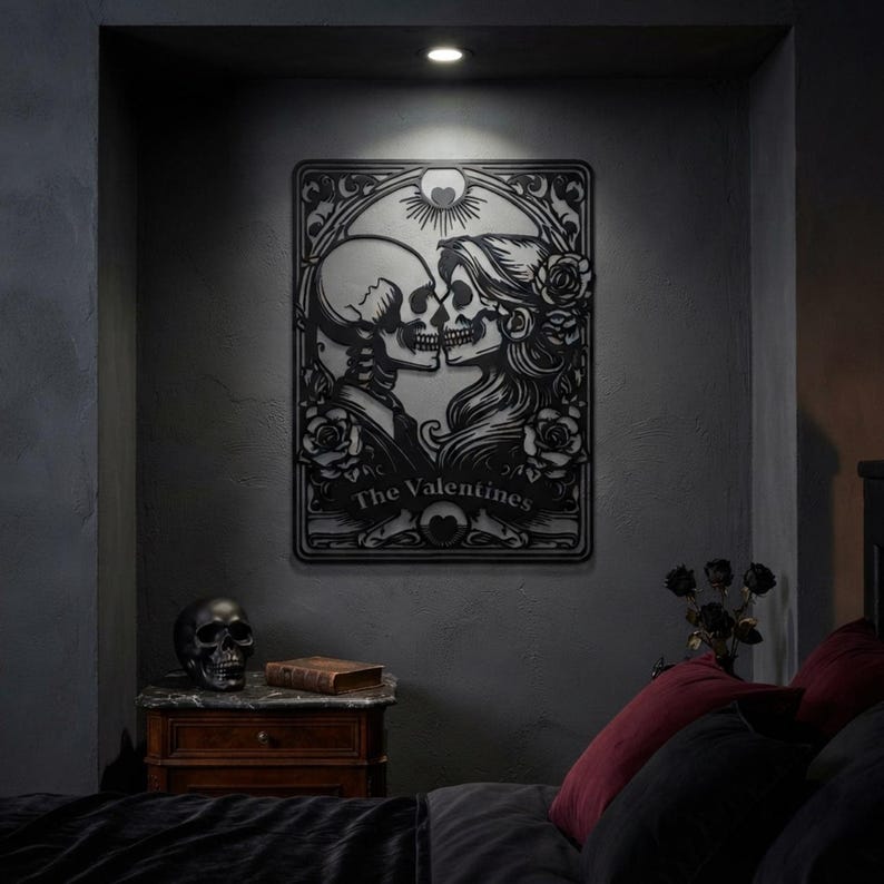 May include: A black metal wall art piece featuring a rectangular frame with a central illustration of two skulls facing each other. The design includes roses, a sunburst motif, and the text The Valentines printed in a banner at the bottom.