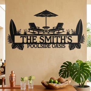 Könnte beinhalten: Ein schwarzes Metall-Wandschild mit einem Scherenschnitt-Design aus zwei Liegestühlen, einem kleinen Tisch und einem Sonnenschirm unter dem Text THE SMITH'S POOLSIDE OASIS. Das Schild ist beidseitig von Surfbrett- und Tropenblatt-Silhouetten flankiert.