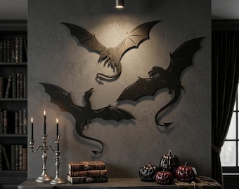Puede incluir: Tres siluetas de dragones de metal negro montadas en una pared gris oscuro. Cada dragón está representado en una pose de vuelo diferente con las alas extendidas y colas largas. El arte de pared se sitúa sobre una repisa con libros y calabazas.