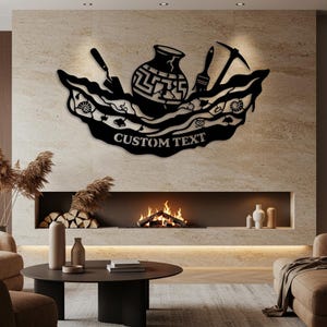 Peut inclure: Décoration murale en métal noir avec un vase central, des outils et le texte "CUSTOM TEXT". L'œuvre comprend une truelle, un pinceau et une pioche, sur un fond décoratif en forme de vague. Idéal pour la décoration intérieure.