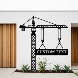 Może przedstawiać: Czarna metalowa grafika ścienna z żurawiem z napisem "CUSTOM TEXT". Żuraw ma wysoką wieżę, poziomy wysięgnik i wiszące haki. Znak jest zawieszony na hakach. Grafika jest zamontowana na białej ścianie.