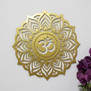 Op de afbeelding: Een goudkleurige, lasergesneden Om-symbool mandala wandkunst. Het ingewikkelde ontwerp heeft een centraal Om-symbool omgeven door een bloemenpatroon. Een tros paarse orchideeën staat in de rechter benedenhoek.