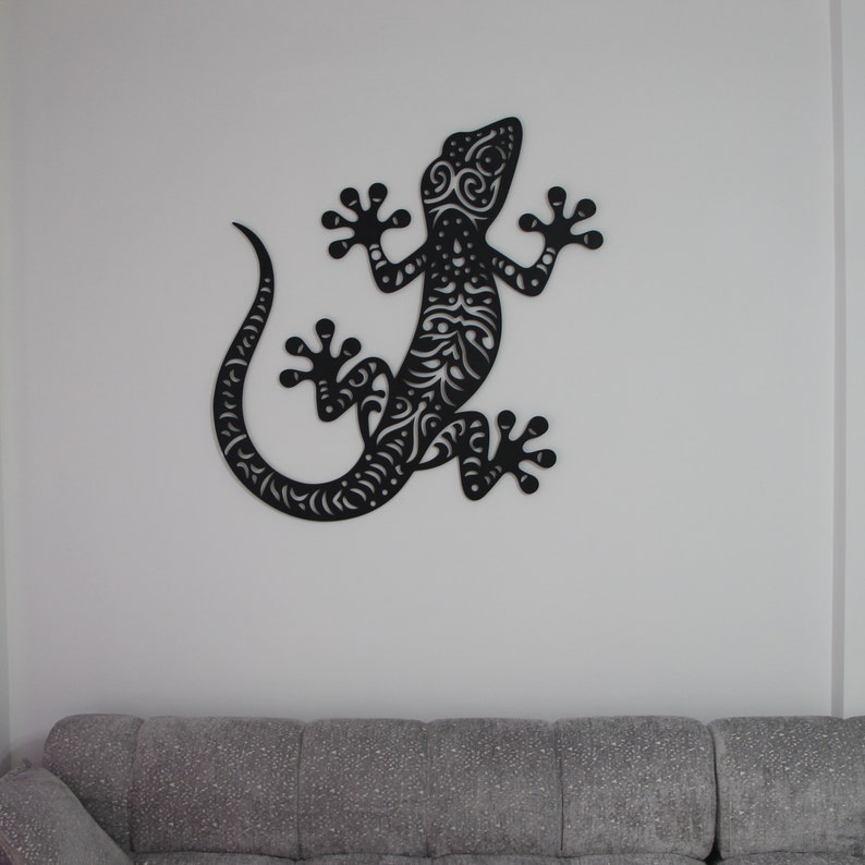 May include: Black wall decor in the shape of a gecko with intricate patterns. The gecko is mounted on a white wall.