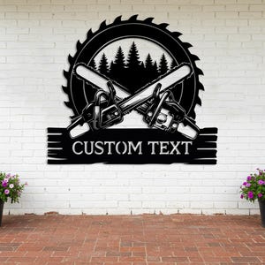 May include: Black metal wall art featuring crossed chainsaws, a circular saw blade, and a forest silhouette. The design includes the text "CUSTOM TEXT" on a rectangular banner. This piece is suitable for a workshop or garage.