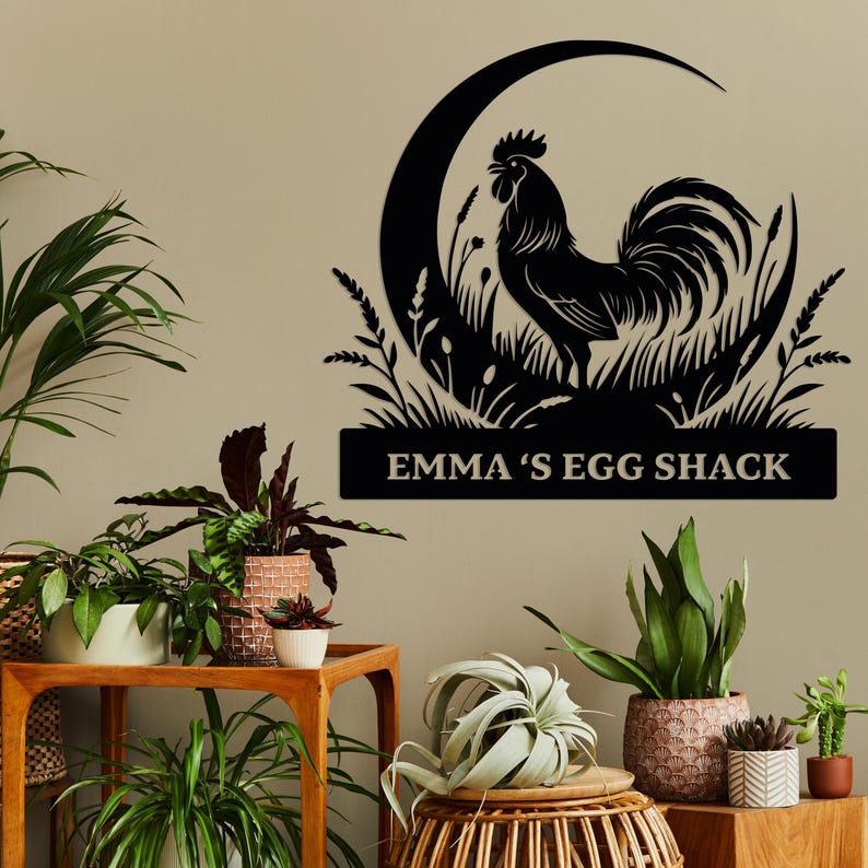 May include: Black metal wall art featuring a rooster silhouette within a crescent moon and grassy design. The text "EMMA'S EGG SHACK" is displayed below. The artwork is mounted on a neutral-colored wall, surrounded by various potted plants.