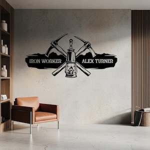 Può includere: Un'opera d'arte da parete in metallo nero raffigurante una lanterna centrale affiancata da due picconi incrociati. Il design include uno stendardo orizzontale con il testo IRON WORKER a sinistra e ALEX TURNER a destra, su una sagoma di montagne.