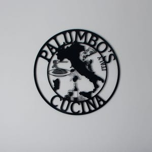 Może przedstawiać: Czarny metalowy znak z napisem "Palumbo's Cucina" w k&oacute;łku. Wewnątrz k&oacute;łka znajduje się sylwetka Włoch z kawałkiem pizzy, talerzem spaghetti, butelką wina i kieliszkiem wina.