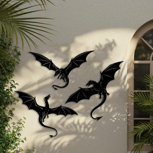 以下が含まれることがあります： 黒い金属製のドラゴンの壁掛けアートが3つ、ベージュの壁に飾られています。 ドラゴンは翼を広げ、詳細な特徴と曲がった尾を持っています。 アートは窓と緑の近くにあります。