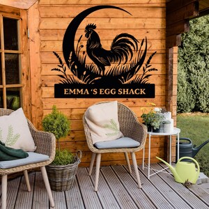 May include: Black metal rooster silhouette wall art. The rooster is set against a crescent moon and tall grass. The text "EMMA'S EGG SHACK" is displayed below the rooster. The art is mounted on a wooden wall.