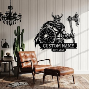 May include: Black metal Viking wall art featuring a warrior with a shield and axe. The design includes the text "CUSTOM NAME EST. 2021". The artwork is mounted on a white wall, with a leather chair and ottoman in the foreground.