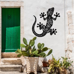 May include: Black metal gecko wall art with intricate details, mounted on a white wall with green plants in pots below.