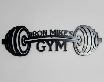 May include: Black metal wall art featuring a barbell with weight plates on each end. The barbell has the text "IRON MIKE'S GYM" written on it.
