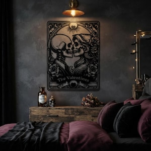 May include: A black metal wall art piece featuring a tarot card design with two skulls facing each other, surrounded by floral patterns and a sun motif. The text "The Valentines" is printed in a banner at the bottom of the rectangular frame.