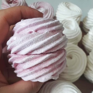 May include: A close-up of a pink and white marshmallow swirl. The marshmallow is held in a hand and is surrounded by other white marshmallows.