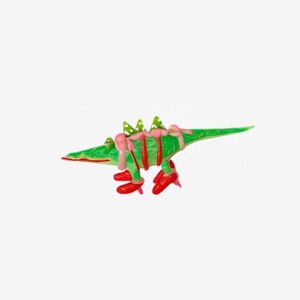 May include: A whimsical green alligator figurine with red stripes and red shoes. The alligator has pink frosting-like accents and green spiky details on its back. The figurine is set against a plain white background.