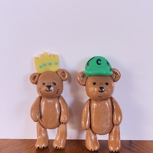 May include: Two brown teddy bears, one wearing a yellow crown and the other wearing a green baseball cap with the letter "C" on it.