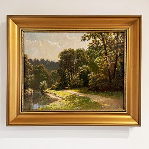 Può includere: Un dipinto ad olio incorniciato che rappresenta una scena di paesaggio con un fiume sinuoso, alberi verdi lussureggianti e un sentiero sterrato che conduce nel bosco. Il dipinto è ambientato su uno sfondo di cielo blu con nuvole bianche.