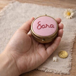May include: A circular, brass-toned keepsake box with a pink fabric top embroidered with the name "Sara" in red thread. The box is held in a hand, with a tooth and a coin nearby. The box has a hinged lid and a decorative border.