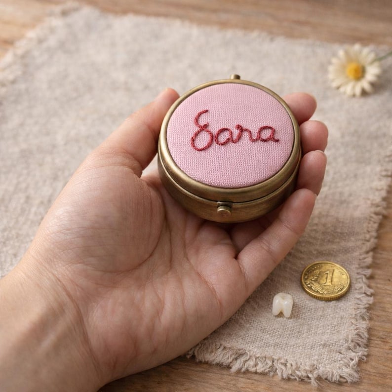 May include: A circular, brass-toned keepsake box with a pink fabric top embroidered with the name "Sara" in red thread. The box is held in a hand, with a tooth and a coin nearby. The box has a hinged lid and a decorative border.