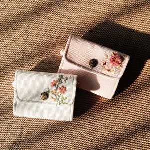Pu&ograve; includere: Due piccoli portafogli in velluto a coste, bianco sporco e rosa pallido, ciascuno ornato da ricami floreali. I portafogli hanno una chiusura a patta con un bottone decorativo. Lo sfondo &egrave; un tessuto beige strutturato.