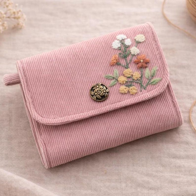 Pu&ograve; includere: Un portafoglio rosa in velluto a coste con un design floreale ricamato. Il portafoglio presenta una chiusura a patta fissata da un bottone decorativo. Il ricamo include fiori bianchi, arancioni e gialli con foglie verdi.