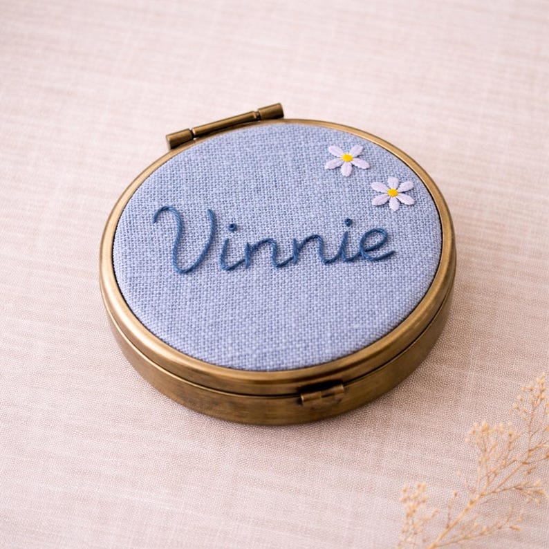 May include: A circular, antique brass compact with a light blue fabric top. The fabric is embroidered with the name "Vinnie" in blue script, and two small white daisies. The compact is closed with a hinged clasp.