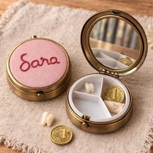 May include: A vintage-style brass tooth fairy box with a pink lid embroidered with the name "Sara" in red thread. The open box reveals a mirror, a tooth, a coin, and a small paper with a heart.