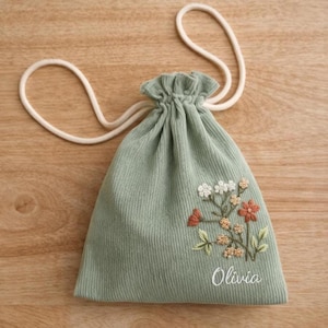 May include: Sage green corduroy drawstring bag with embroidered floral design in white, orange, and tan. The bag has a white drawstring and the name "Olivia" embroidered on the front.