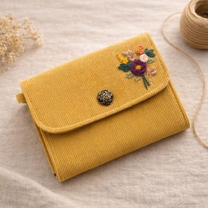 Pu&ograve; includere: Portafoglio in velluto a coste giallo senape con ricamo floreale. Il portafoglio ha una chiusura con patta con un bottone decorativo. I fiori ricamati includono accenti viola, gialli e verdi. Sul lato c'&egrave; un piccolo anello.