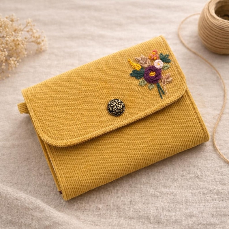 Pu&ograve; includere: Portafoglio in velluto a coste giallo senape con ricamo floreale. Il portafoglio ha una chiusura con patta con un bottone decorativo. I fiori ricamati includono accenti viola, gialli e verdi. Sul lato c'&egrave; un piccolo anello.