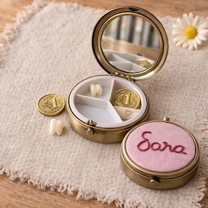 May include: A circular brass keepsake box with a mirror, open to reveal compartments containing a tooth, a coin, and a ribbon. A second box with the name "Sara" embroidered on a pink lid is also visible. The boxes are on a beige cloth.