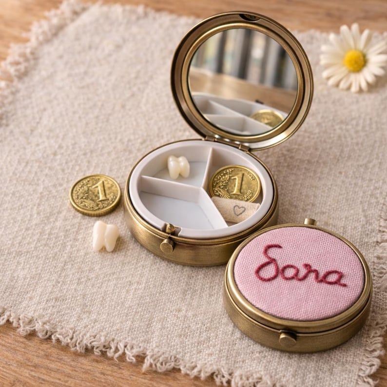 May include: A circular brass keepsake box with a mirror, open to reveal compartments containing a tooth, a coin, and a ribbon. A second box with the name "Sara" embroidered on a pink lid is also visible. The boxes are on a beige cloth.