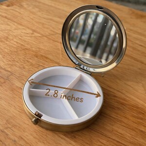 May include: Open, round, gold-toned pill box with a mirror and a white interior divided into three compartments. The box is 7.1 cm in diameter. The pill box is on a wooden surface.