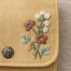 Pu&ograve; includere: Un oggetto in tessuto di velluto a coste giallo con un ricamo floreale. Il ricamo presenta fiori e foglie marroni, bianchi e verde chiaro. Un bottone decorativo in metallo con un motivo floreale &egrave; sul lato sinistro.