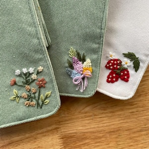 Pu&ograve; includere: Primo piano di tre articoli in tessuto ricamato. Due sono verde salvia, uno &egrave; bianco. Gli articoli verdi presentano ricami floreali, uno con un bouquet e l'altro con una variet&agrave; di fiori. L'articolo bianco ha fragole e fiori ricamati.