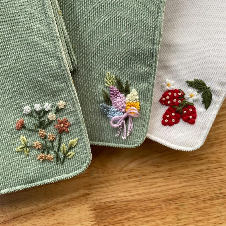 Pu&ograve; includere: Primo piano di tre articoli in tessuto ricamato. Due sono verde salvia, uno &egrave; bianco. Gli articoli verdi presentano ricami floreali, uno con un bouquet e l'altro con una variet&agrave; di fiori. L'articolo bianco ha fragole e fiori ricamati.