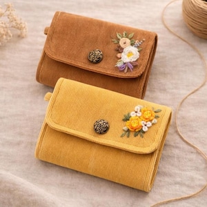 Pu&ograve; includere: Due portafogli in velluto a coste, uno marrone caldo e l'altro giallo dorato. Ogni portafoglio presenta un ricamo floreale e una chiusura con bottone decorativo. I portafogli sono piccoli e rettangolari, con chiusura a patta.