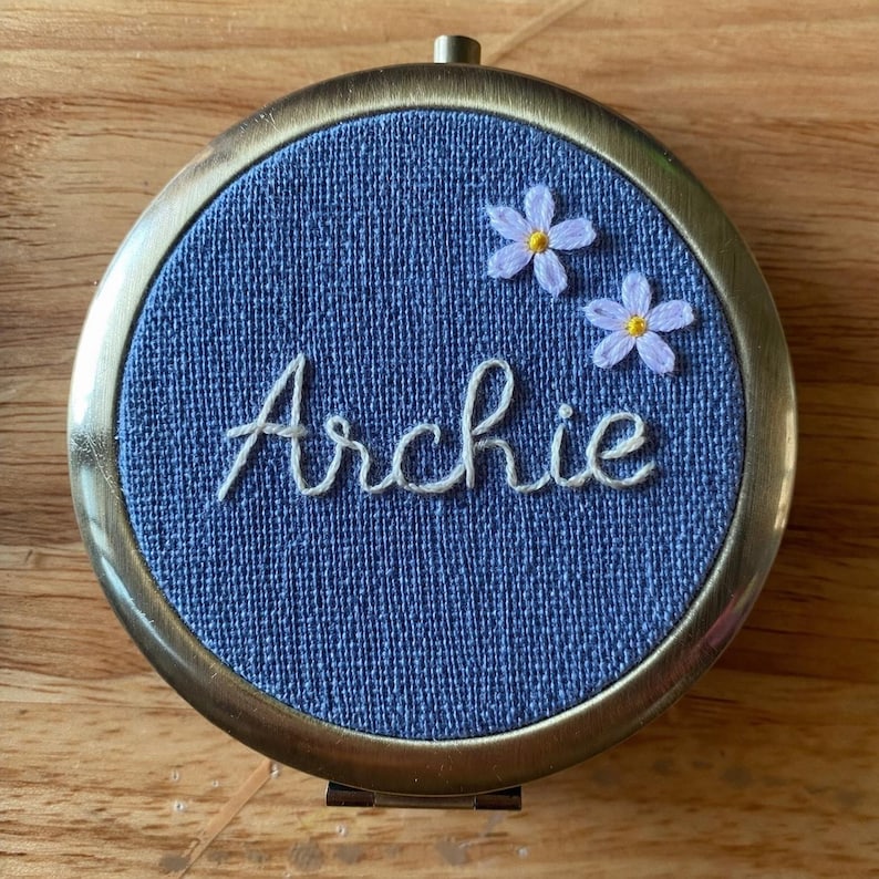 May include: A circular, antique brass compact mirror with a blue fabric insert. The insert features the name "Archie" embroidered in white script, and two small embroidered lavender flowers. The compact has a hinged opening and a small loop at the top.
