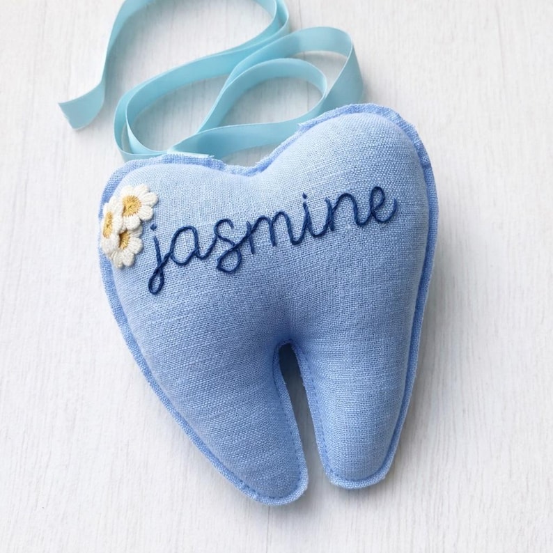 Może przedstawiać: Jasnoniebieska poduszka w kształcie zęba z wyhaftowanym imieniem "jasmine" ciemnoniebieskim pismem. Mała kępka białych i ż&oacute;łtych kwiat&oacute;w jest przyszyta po lewej stronie. Do zawieszenia przymocowana jest jasnoniebieska wstążka.