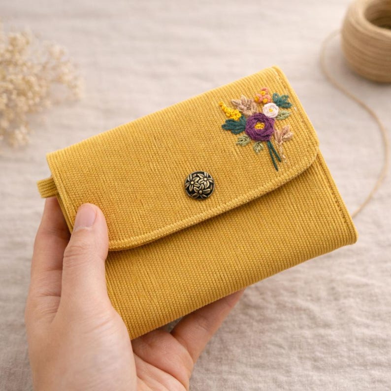 Pu&ograve; includere: Portafoglio in velluto a coste giallo senape con ricamo floreale. Il portafoglio ha una chiusura con patta e un bottone decorativo. I fiori ricamati includono fiori viola, gialli e bianchi con foglie verdi.