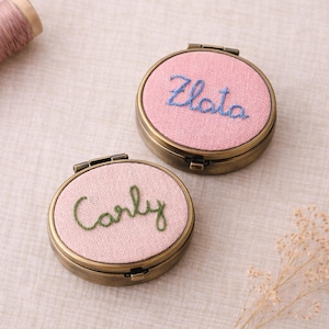 May include: Two small, round, antique brass compact mirrors. Each has a pink fabric centre with embroidered names: "Zlata" in blue and "Carly" in green. The mirrors are open, revealing the interior.
