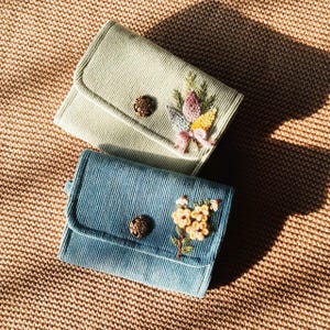 Pu&ograve; includere: Due portafogli in velluto a coste, uno verde chiaro e uno blu, con ricami floreali e chiusure con bottoni. I portafogli sono rettangolari con patta e sono su una superficie marrone strutturata.