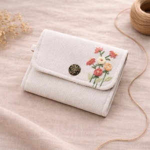 Pu&ograve; includere: Un portafoglio in velluto a coste beige chiaro con un design ricamato floreale. Il portafoglio presenta una chiusura con bottone in metallo decorativo e una chiusura a patta. I fiori ricamati includono sfumature di rosa, arancione e crema, con foglie verdi.