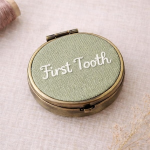 May include: A circular, antique-style metal box with a hinged lid. The top features light green fabric embroidered with the words "First Tooth" in white. This box is designed to store a baby's first tooth.