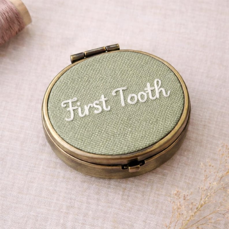 May include: A circular, antique-style metal box with a hinged lid. The top features light green fabric embroidered with the words "First Tooth" in white. This box is designed to store a baby's first tooth.