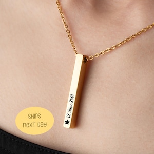 Könnte beinhalten: Eine goldfarbene Halskette mit Anhänger in Form eines Stabes. Der rechteckige Anhänger trägt den eingravierten Text "12. Juni 2001" mit einem Sternsymbol. Die Halskette ist ebenfalls goldfarben. Die Halskette wird um den Hals getragen.