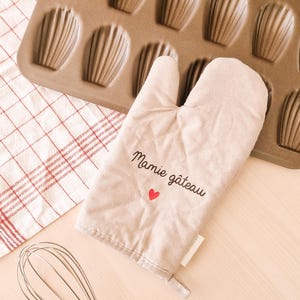 Peut inclure: Un gant de cuisine beige avec le texte "Mamie gâteau" et un cœur rouge brodé dessus. Le gant est posé sur une surface en bois à côté d'un fouet en métal et d'un moule à pâtisserie avec 12 empreintes individuelles.