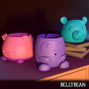 Puede incluir: Tres macetas de cerámica de colores con forma de cerdo, panda y oso. La maceta de cerdo es rosa, la maceta de panda es morada y la maceta de oso es azul. Las macetas están sobre una superficie de madera.